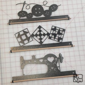 May include: Three metal wall-mounted fabric storage rods with decorative cutouts. The top rod features a needle and thread, buttons, and a loop. The middle rod has three quilt block designs. The bottom rod has a sewing machine design.
