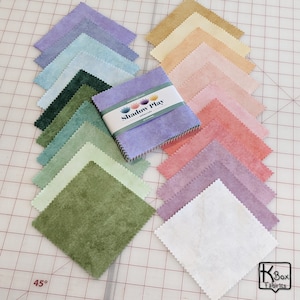 May include: A collection of square fabric swatches in various pastel colors, including lavender, green, and peach. A bundle of fabric squares is wrapped with a label that reads "Shadow Play Minerals". The fabric squares are arranged on a grid.