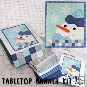 May include: A tabletop banner kit featuring a snowman design. The kit includes a finished quilt, fabric squares, and a box with the snowman image. The snowman has a carrot nose, black eyes, and a blue hat. The text "TABLETOP BANNER KIT" is visible.