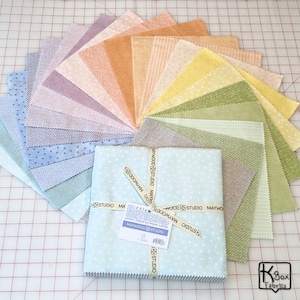 May include: A collection of fabric squares in various pastel colors, arranged in a fan shape. The fabrics have different patterns, including dots, stripes, and textures. A stack of light blue fabric with white polka dots is tied with a ribbon that says "MAYWOOD STUDIO".