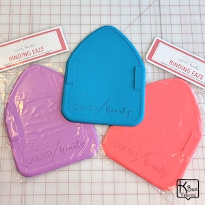 May include: Three silicone binding ease tools in different colors: purple, blue, and orange. Each tool has the text "QUILTED/heartz" embossed on it.