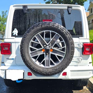 以下が含まれることがあります： スペアタイヤ付きの白いSUVの後部ビュー。タイヤは黒とシルバーのリムで、オレンジ色のアクセントが付いています。小さなイエティのステッカーがリアウィンドウに貼られています。赤いテールライトと青い牽引フックが見えます。