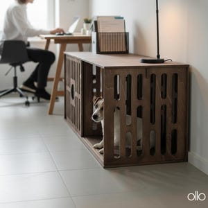以下が含まれることがあります： 濃い色の木製犬用クレートで、スロットデザインが特徴です。小さな犬がクレートの中にいます。クレートは、黒いデスクランプと、背景でラップトップで作業している人がいるデスクセットアップの一部です。