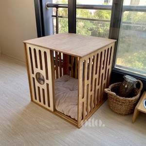 May include: A wooden pet house with a light brown finish. The house has a square shape with cut-out vertical and horizontal lines on the sides. Inside is a beige pet bed. The house is near a window and a woven basket.
