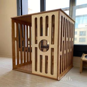 May include: A wooden pet crate with a sliding door and vertical cutouts. The crate has a natural wood finish and a circular cutout with the word "otto" visible. The crate is designed for indoor use.