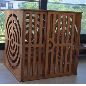 May include: A wooden cat playhouse with a circular cutout pattern on one side and vertical slats on the other sides. The playhouse is made of light brown wood and has a square shape.