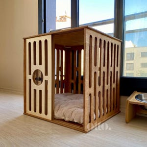 May include: Wooden dog crate with a light brown finish. The crate features a sliding door and vertical slats for ventilation. The interior includes a plush, beige bed. The word "otto" is visible on the front. A matching food bowl stand is next to the crate.