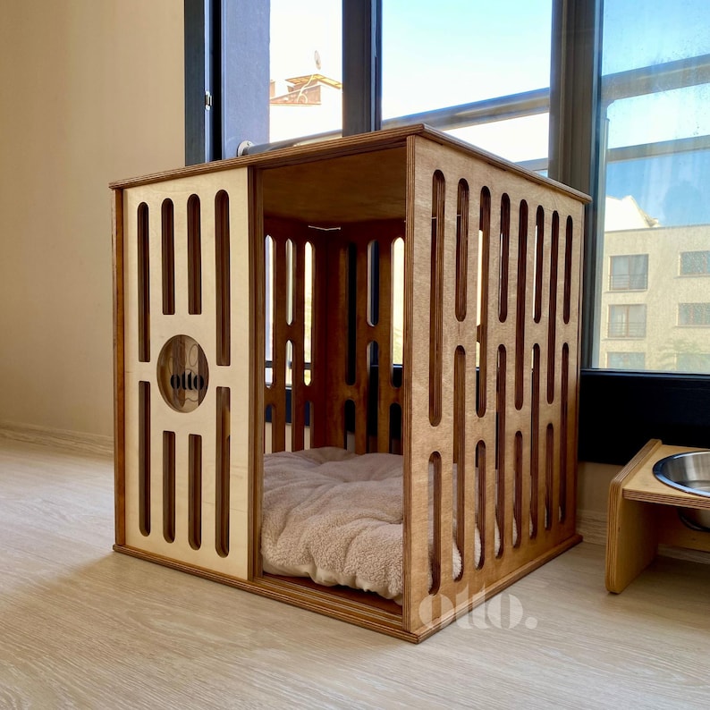 May include: Wooden dog crate with a light brown finish. The crate features a sliding door and vertical slats for ventilation. The interior includes a plush, beige bed. The word "otto" is visible on the front. A matching food bowl stand is next to the crate.