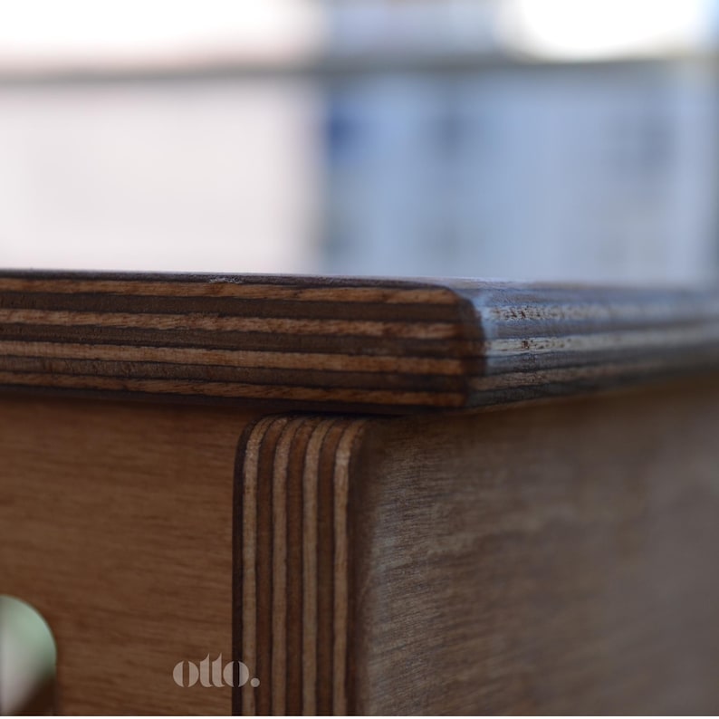 May include: Close-up of the top edge of a wooden table with a dark brown stain. The table top is made of plywood with visible layers of wood veneer. The edge of the table is rounded and smooth.