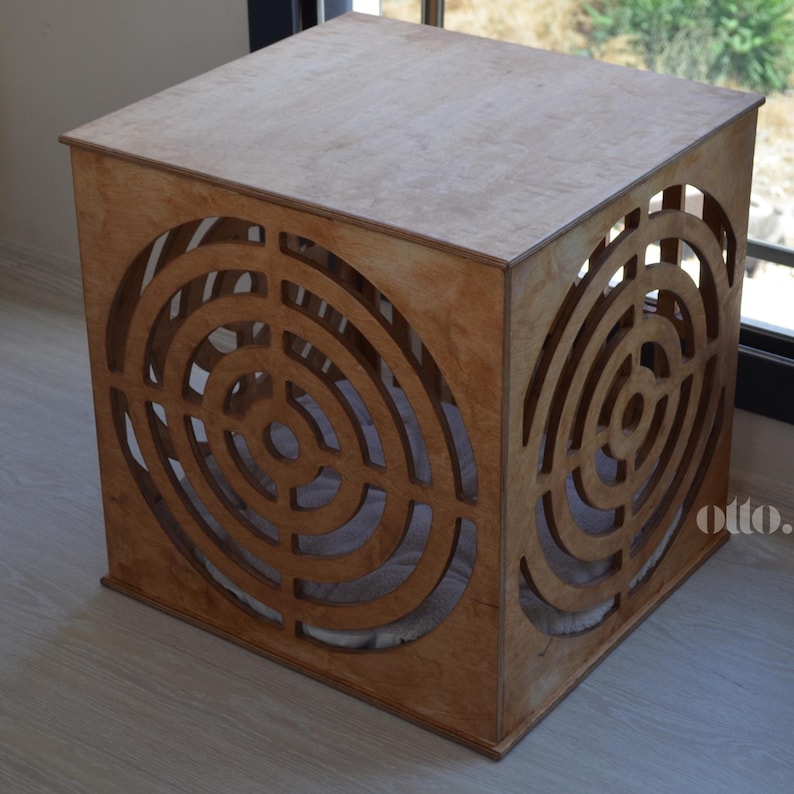 May include: A wooden cube pet bed with a circular cutout pattern on each side. The bed is made of light brown wood and has a natural finish. The bed is sitting on a light brown floor.