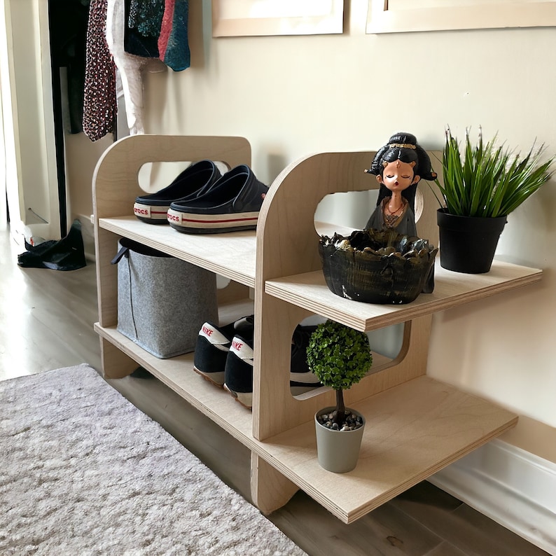 May include: A light wood shelf unit with three shelves. The top shelf has a small potted plant, a decorative bowl, and a doll. The middle shelf has a small wicker basket and the bottom shelf has two pairs of shoes.