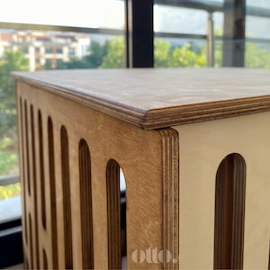 May include: A light-colored wooden cube-shaped storage unit with rounded cutouts on the sides. The top and sides are made of layered wood, with a natural finish. The word "otto!" is visible on the lower right side.