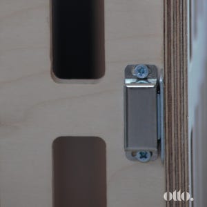 May include: Close-up of a silver magnetic cabinet door catch mounted on a light wood surface. The catch has two screws visible.
