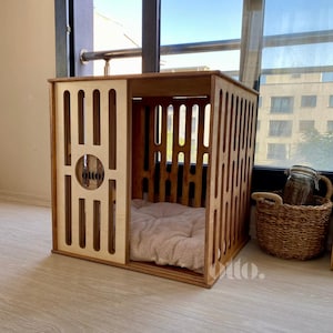 May include: A wooden dog crate with a light brown finish. The crate has a rectangular shape with vertical slats on the sides and a circular design with the word "otto" on the front. Inside is a soft, beige pet bed.