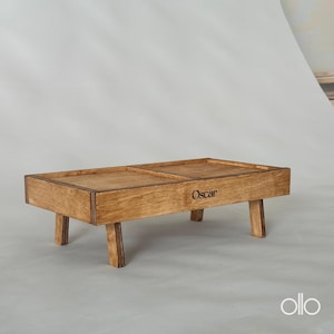 Könnte beinhalten: Ein rechteckiges Serviertablett aus Holz mit vier kurzen Beinen. Das Tablett ist in zwei Abschnitte unterteilt und trägt den Namen "Oscar" auf der Vorderseite eingraviert. Das Holz hat eine natürliche, hellbraune Oberfläche.