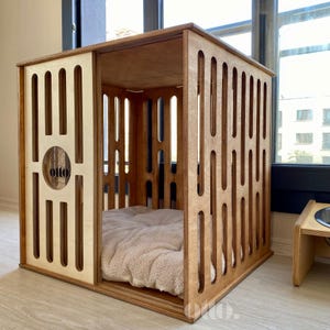 May include: A wooden dog house with a square design. The structure features vertical and curved cutouts on the sides and a sliding door. The interior includes a plush, light-colored bed. The word "otto" is visible on the front.