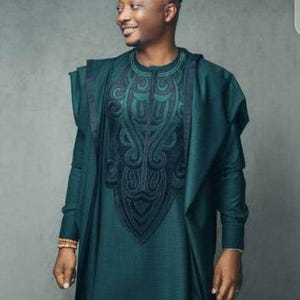 May include: A teal green traditional outfit with intricate black embroidery on the chest. The ensemble includes a matching hat and a draped shawl. The outfit is worn by a person smiling, suggesting a celebratory occasion.