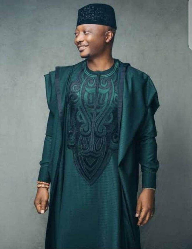 May include: A teal green traditional outfit with intricate black embroidery on the chest. The ensemble includes a matching hat and a draped shawl. The outfit is worn by a person smiling, suggesting a celebratory occasion.