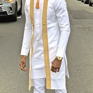 May include: White tunic and trousers set with gold detailing. The tunic has gold trim and a tassel. The outfit is paired with sandals. The person is standing on a street.