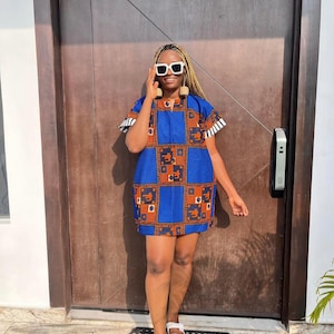 May include: A woman wearing a blue and orange patterned short-sleeved dress with white accents. She is wearing white sunglasses and white sandals. She is standing in front of a wooden door.
