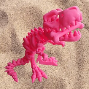Puede incluir: Un juguete de esqueleto de dinosaurio de plástico rosa. El dinosaurio está en posición de pie con la boca abierta y la cabeza inclinada hacia un lado. El juguete está hecho de plástico rosa brillante y tiene una estructura esquelética detallada.