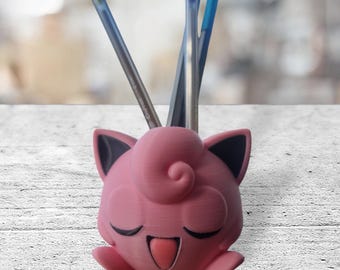 May include: Pink 3D printed Pokemon Jigglypuff pencil holder with black ears and eyes.  The holder has a small pink tongue sticking out and is holding several blue and silver pencils.