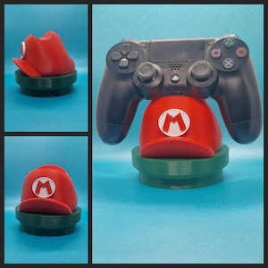 May include: A black video game controller is sitting on a red and green Mario hat-shaped stand. The stand is shaped like a red cap with a white 'M' on the front and a green base.