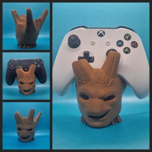 May include: A brown, wooden, Groot-inspired figure with a smiling face, holding a white Xbox controller. The figure is a stand for a video game controller.