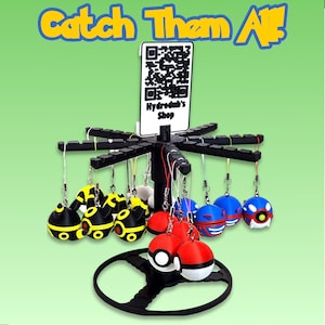 May include: A black display stand holding various colorful character-themed keychains. The keychains are round and feature designs in yellow, red, blue, and white. The stand has a sign with a QR code and the text "Hydrodub's Shop" and the words "Catch Them All!" in yellow and blue.