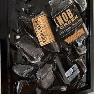 May include: A framed artwork featuring shattered glass and fragments of Knob Creek bourbon bottles. The labels are visible, with text including "Single Barrel Select" and "Kentucky Straight Bourbon Whiskey". The artwork is set against a black background.