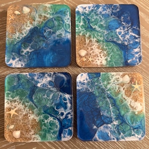 Puede incluir: Conjunto de cuatro posavasos cuadrados con temática de playa. Cada posavasos presenta un diseño abstracto azul, verde y blanco con purpurina dorada y conchas blancas.