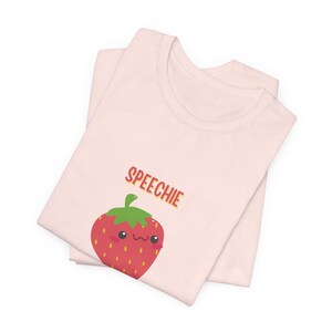 May include: Two light pink t-shirts are folded and stacked. The top shirt features a red strawberry graphic with a smiling face and the word "SPEECHIE" in orange and red letters.