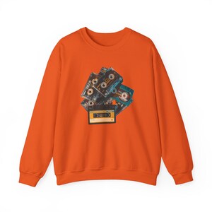 May include: Orange sweatshirt featuring a graphic of several vintage cassette tapes in shades of black, blue, and yellow. The cassette tapes are arranged in a pile, with details like labels and tape reels visible. The sweatshirt has a crew neck and long sleeves.