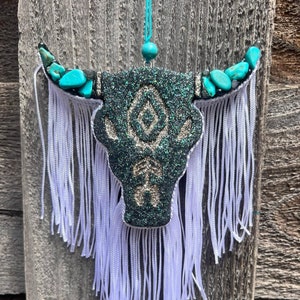 May include: A decorative bull skull air freshener with turquoise accents on the horns and a teal glitter design. White fringe hangs below the skull. The text "fancyfreshies.shop" is at the bottom.