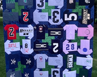 May include: A blue and green patchwork quilt made from t-shirts with numbers, logos, and text. The quilt features a variety of sports teams, including Seattle Volleyball, Bryant Volleyball, and EWHS Volleyball. The quilt is made up of squares and rectangles, and the seams are visible.