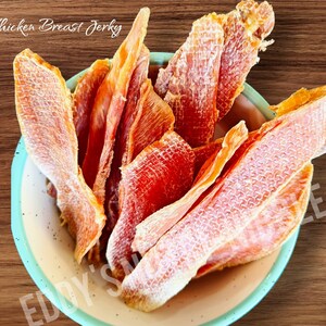May include: A bowl filled with chicken breast jerky. The jerky is a reddish-brown color and has a textured surface. The bowl is beige with a light blue rim. The words "Chicken Breast Jerky" are written in cursive at the top of the image.