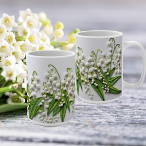 Può includere: Due tazze in ceramica bianca con un motivo floreale di fiori di mughetto bianchi e foglie verdi. Le tazze sono su una superficie di legno.