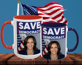May include: Two white ceramic mugs with red and blue rims. Both mugs feature a photo of Kamala Harris and the text "Save Democracy". The mugs are on a wooden surface with an American flag in the background.