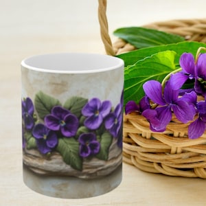 May include: A white ceramic mug with a design of purple violets on a wooden background. The mug is sitting on a wooden table next to a wicker basket with purple flowers.