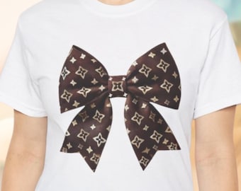 May include: A white t-shirt with a brown bow printed on the front. The bow is made of a fabric with a brown and gold pattern of small four-pointed stars.