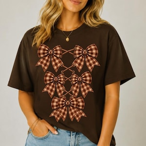 Może przedstawiać: Ciemnobrązowy t-shirt z krótkim rękawem z nadrukiem graficznym przedstawiającym pięć brązowo-białych kokard w kratę ułożonych w pionowy wzór. Kokardy są połączone cienkimi, krzyżującymi się liniami, tworząc strukturalny wzór na przodzie koszulki.