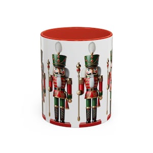 Könnte beinhalten: Eine weiße Keramiktasse mit rotem Rand zeigt ein sich wiederholendes Muster von Nussknacker-Soldaten. Die Nussknacker tragen rot-grüne Uniformen mit goldenen Akzenten und halten einen goldenen Stab.
