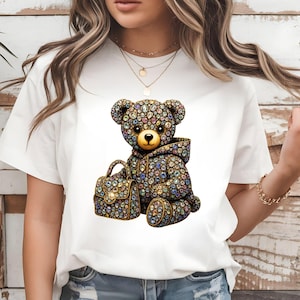May include: White t-shirt with a jewelled teddy bear design. The bear wears a hoodie and carries a matching purse, both covered in colourful gemstones. The purse has a gold clasp. The design is centered on the shirt.