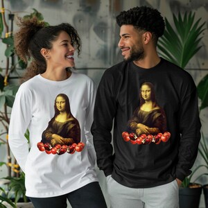 Puede incluir: Dos personas con camisas de manga larga con un estampado gráfico de la Mona Lisa sosteniendo manzanas. La mujer lleva una camisa blanca y el hombre lleva una camisa negra.