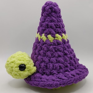 May include: A crocheted purple and green turtle wearing a purple witch hat with a green band. The hat is pointy and the turtle has a small, black eye.
