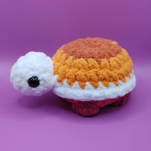 May include: A plush, crocheted turtle toy. The turtle's shell is a gradient of orange and brown, outlined in white. The head is white with a black eye, and the feet are a deep pink. The toy is set against a purple background.