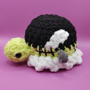 May include: A handmade crochet turtle toy. The turtle has a black shell, a light green head, and a white and green body. The shell has a stitched design. The turtle's eye is a black bead. The toy is made of soft, fluffy yarn.