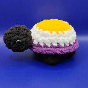 May include: A handmade crochet turtle in black, purple, white, and yellow. The turtle's shell features a non-binary pride flag design. The turtle has a black head with a black eye and a black body.