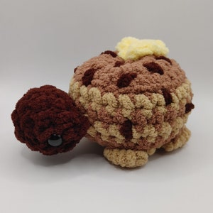 May include: A crocheted turtle toy with a brown head and legs, a tan shell with brown spots, and a yellow flower on top.