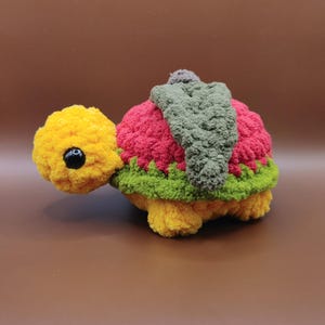 May include: A handmade, plush turtle toy with a yellow head and feet, a red and green shell, and a gray leaf-like detail on its back. The turtle has a black eye and is made of soft, textured yarn.